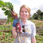 A young woman with blond hair standing in a farm field holding recently picked beets