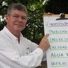 A man in a white Shirt is pointing to a flipchart showing a list of community challenges identified at a community needs assessment.