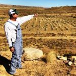 Hopi dry farmer with blue overalls and a white hat pointing toward his dry, brown farm fields that extend into the distance